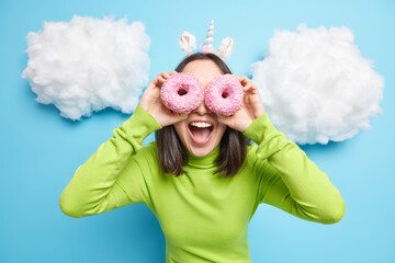Positive Asian woman covers eyes with two glazed doughnuts keeps mouth opened dressed in green...