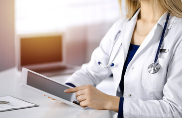 Unknown young woman-doctor is checking some medication names, while sitting at the desk in her sunny cabinet in a clinic. Physician with a stethoscope is using a tablet computer, close-up. Perfect