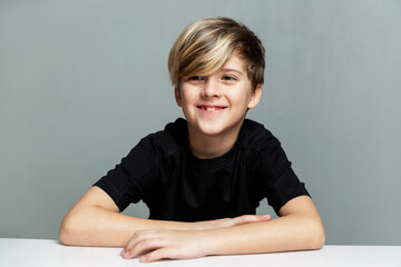 A boy in a black T-shirt with a fashionable hairstyle sits at a table and laughs. Grey background. © Анна Демидова