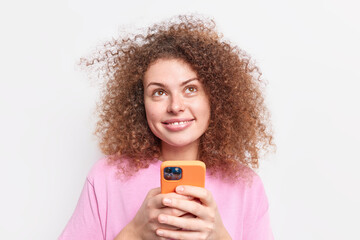 Portrait of good looking young European woman with curly hair holds smartphone concentrated above smiles gently uses social media app chats online dressed casually isolated over white background