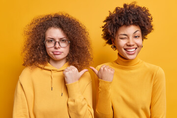 Cheerful Afro American woman and her sad curly haired sister point thumbs at each other express...