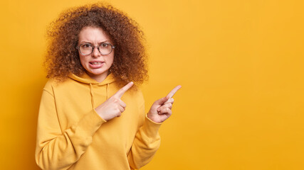 Nervous displeased curly haired woman frowns face says its unfair points at upper right corner with...