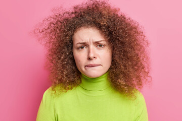 Photo of frustrated unhappy woman with curly hair bites lips looks nervously at camera feels...