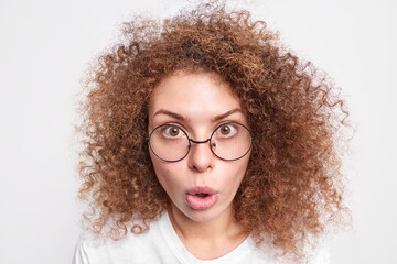 Obraz premium Headshot of surprised European woman with curly bushy hair looks with amazement at camera cannot believe in shocking news wears round spectacles isolated over white studio background. Omg concept