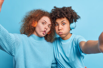 Two mixed race female friends make funny grimaces blow cheeks and fish lips listen music via stereo wireless headphones stand closely to eath other take selfie isolated over blue background.