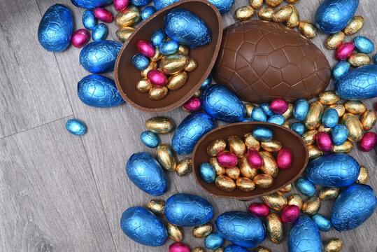 Group of different sizes of colourful foil wrapped chocolate easter eggs in pink, blue, and gold. Large halves of a brown milk chocolate egg have mini eggs inside, on a grey wooden background. - Powered by Adobe