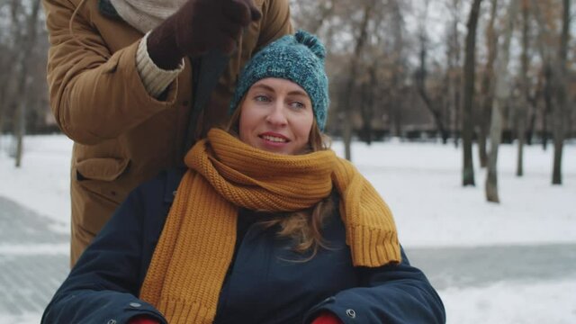 Young positive caucasian woman on wheelchair smiling and discussing something with Afro-American male friend while walking together outdoors on winter day