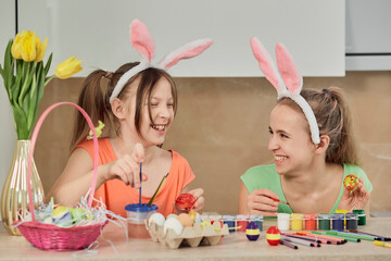 Two girls paint Easter eggs with colorful paint in the kitchen and laugh. Easter holiday.