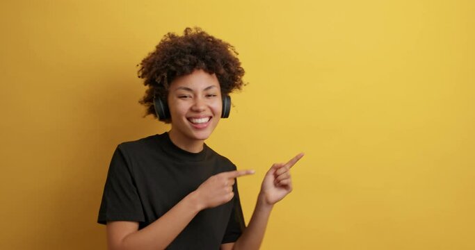 Smiling African American woman shows good deal offer indicates at copy space with good mood listens music via headphones shows advertisement or logo banner makes announcement dressed casually