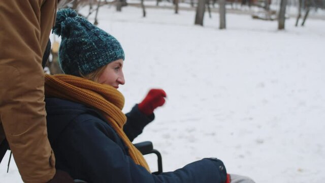 Tilt up shot of young caucasian woman riding wheelchair with help of Afro-American male friend and talking with him while walking outdoors on winter day
