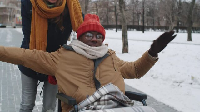 Tilt up shot of joyous Afro-American man on wheelchair smiling and holding arms outstretched while walking with assistance of Caucasian female friend outdoors on winter day