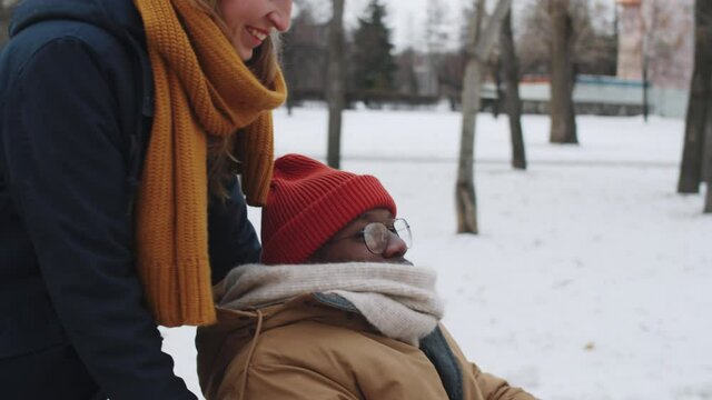Young caucasian woman pushing Afro-American male friend on wheelchair and discussing something with him while walking outdoors on winter day