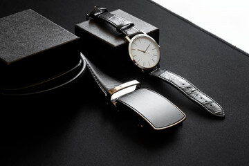 A black leather belt with automatic buckle and a wristwatch lie on a dark surface. The concept of...