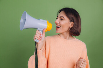 Young woman in casual peach sweater isolated on green olive color background happy funny screaming in megaphone