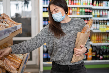 Young woman in a protective mask in the store buys fresh hot bread in the department. Shopping for...