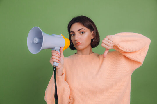 Young woman in casual peach sweater isolated on green olive color background unhappy serious with megaphone shows thumb down