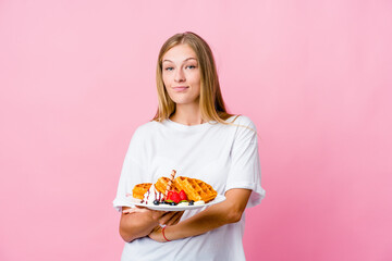 Young russian woman eating a waffle isolated who feels confident, crossing arms with determination.