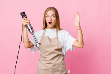 Young russian cook woman holding an electric mixer isolated receiving a pleasant surprise, excited and raising hands.