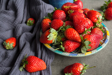 A lot of fresh strawberries on a table. Close up photo of blue ceramic plate and cup, juicy berries and navy blue kitchen towel. Grey concrete background. 
