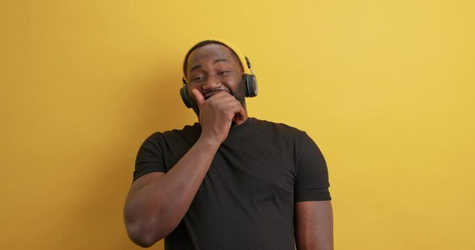 Overjoyed bearded dark skinned guy laughs out hears funny story keeps hand on chest cannot stop laughing listens music via wireless headphones poses against vivid yellow background. Joy concept
