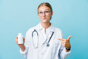 Young russian doctor woman holding pills bottle on blue person pointing by hand to a shirt copy space, proud and confident