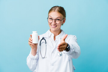 Young russian doctor woman holding pills bottle on blue stretching hand at camera in greeting gesture.