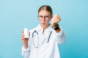 Young russian doctor woman holding pills bottle on blue showing fist to camera, aggressive facial expression.
