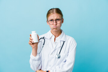 Young russian doctor woman holding pills bottle on blue blows cheeks, has tired expression. Facial expression concept.