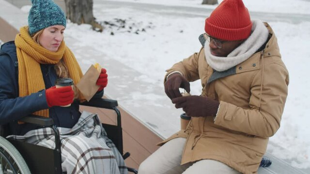 Young paraplegic woman on wheelchair and Afro-American man eating sandwiches, drinking to go coffee, smiling and talking while sitting in park on winter day