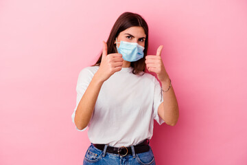 Young caucasian woman wearing a mask for virus isolated on pink background with thumbs ups, cheers about something, support and respect concept.