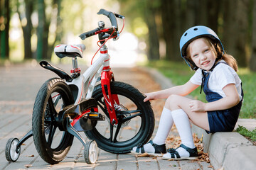 Cheerful smiling girl in helmet sitting near bicycle. Kid checking bike tires