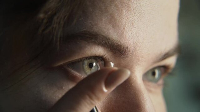 Slow motion macro of green or gray eye of a girl. A finger gently places a dirty contact lens over the iris of the eye. Unsuccessful erroneous insertion of the lens. The girl straightens the lens