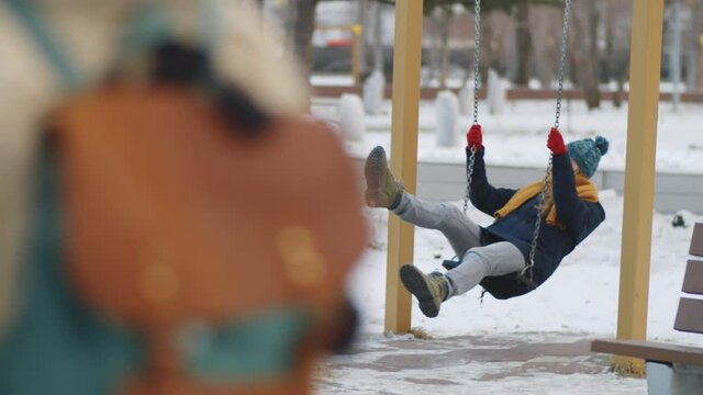 Positive young Caucasian woman swinging on swing, laughing and chatting with disabled male Afro-American friend on wheelchair during walk in park on winter day