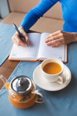 Woman have a teabreak during working hours and planning next work day. Girl writes her plans in a notebook and drinks vitaminic herbal healthy tea from orange and berry sea buckthorn, sitting in cafe