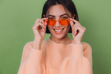 Young stylish woman in casual peach sweater and orange glasses isolated on green olive background happy positive smiling laugh around copy space