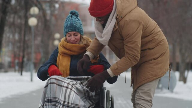 Afro-American man fixing blanket and pushing young cheerful woman on wheelchair while walking in park on winter day
