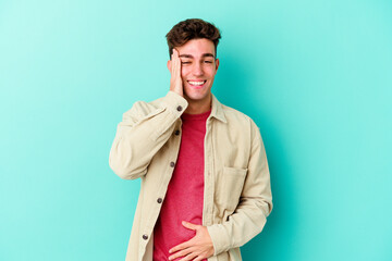 Young caucasian man isolated on blue background laughs happily and has fun keeping hands on stomach.
