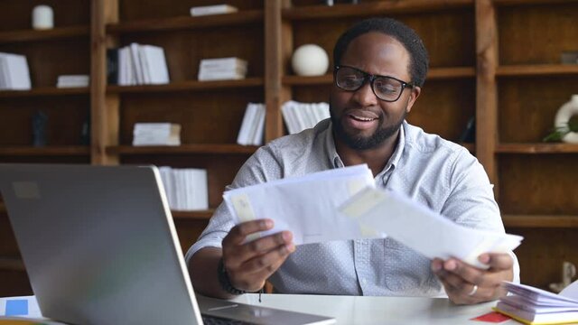 Worried African-American looking through correspondence, a guy holds envelopes in hands, got fine by post, concentrated man sitting at the desk with a laptop in contemporary office