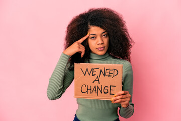 Young african american curly woman holding a we need a change cardboard pointing temple with finger, thinking, focused on a task.