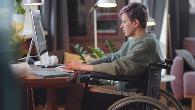 Side view of young disabled woman on wheelchair using computer at desk in living room at home