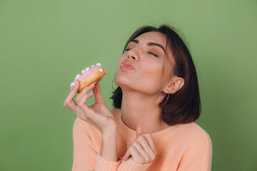 Young stylish woman in casual peach sweater and orange glasses isolated on green olive background biting pink donut with closed eyes copy space