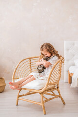 little girl in a white dress sits on a chair and hugs an Easter bunny. A child in a bright room with a toy rabbit.