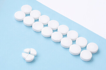 Round white medical pills arranged and one of them is crushed into four parts on blue background. Pill splitting.