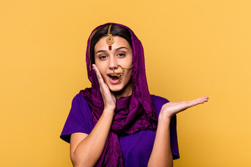 Young Indian woman wearing a traditional sari clothes isolated on yellow background holds copy space on a palm, keep hand over cheek. Amazed and delighted.