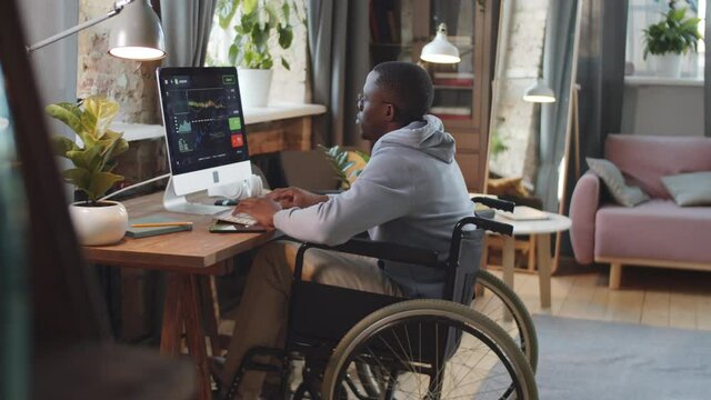 Disabled Afro-American man on wheelchair using computer at desk in living room while working remotely from home