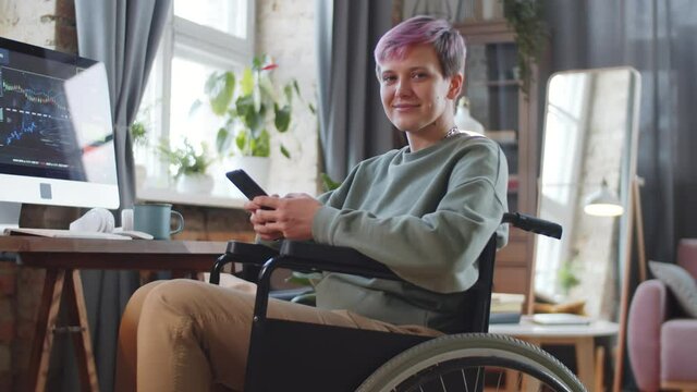Young cheerful disabled woman on wheelchair holding smartphone, looking at camera and smiling while working remotely from home