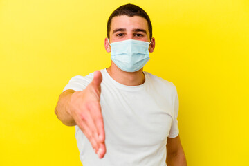 Young caucasian man wearing a protection for coronavirus isolated on yellow background stretching hand at camera in greeting gesture.