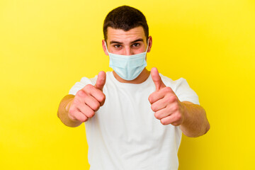 Young caucasian man wearing a protection for coronavirus isolated on yellow background with thumbs ups, cheers about something, support and respect concept.