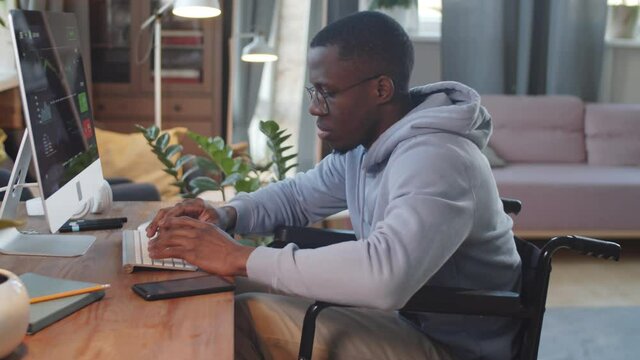 Side view of young Afro-American man on wheelchair typing on computer at desk in living room while working online from home