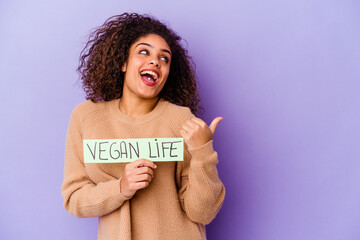 Young African American woman holding a Vegan life placard isolated points with thumb finger away, laughing and carefree.